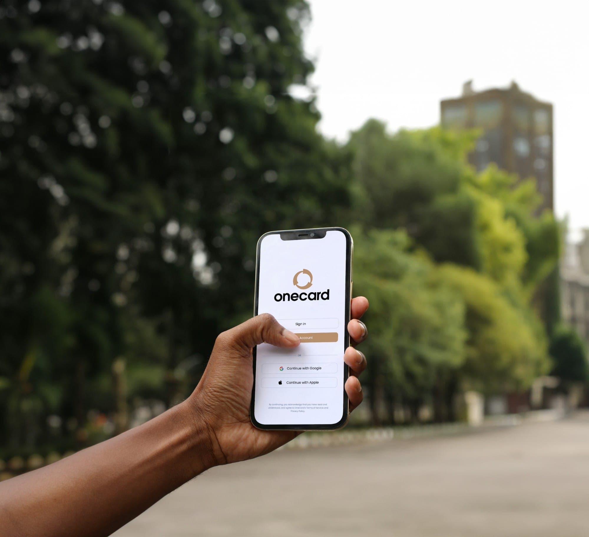 A hand holding up a phone showing the OneCard sign-in screen, trees behind